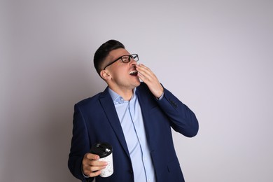 Tired businessman with mug of drink yawning on light grey background