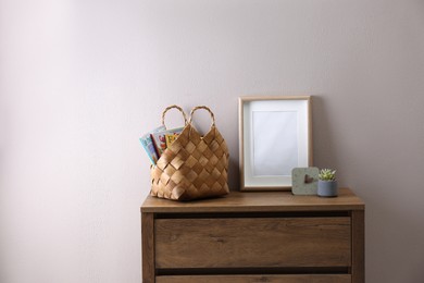 Wooden chest of drawers with stylish bag, decor and empty frame near light wall in room. Interior design