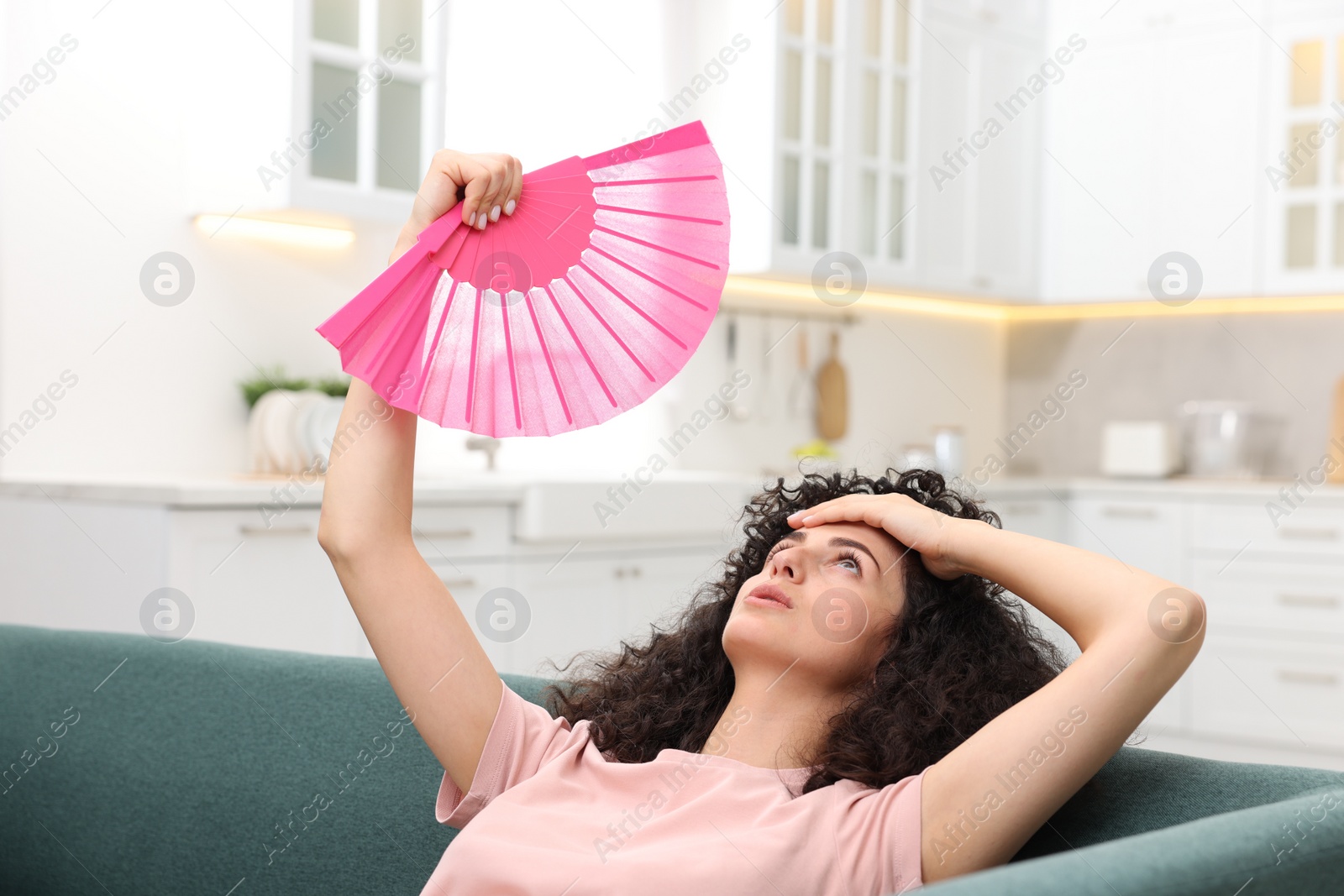 Photo of Young woman waving pink hand fan to cool herself on sofa at home