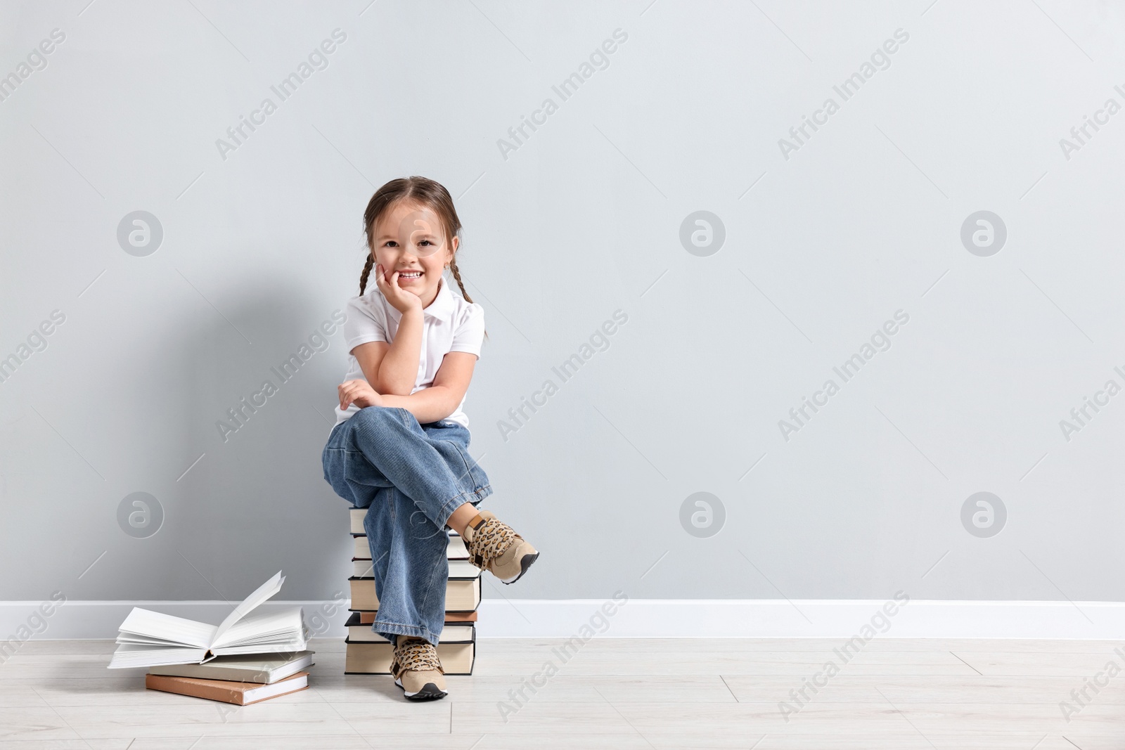 Photo of Cute little girl sitting on stack of books near light grey wall. Space for text