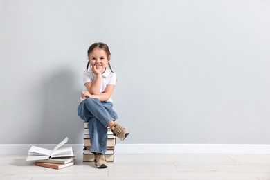 Photo of Cute little girl sitting on stack of books near light grey wall. Space for text