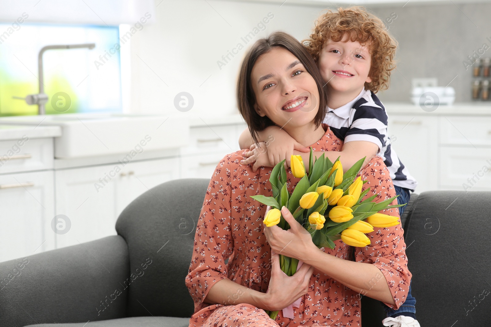 Photo of Little son congratulating his mom with Mother`s day at home, space for text. Woman holding bouquet of yellow tulips