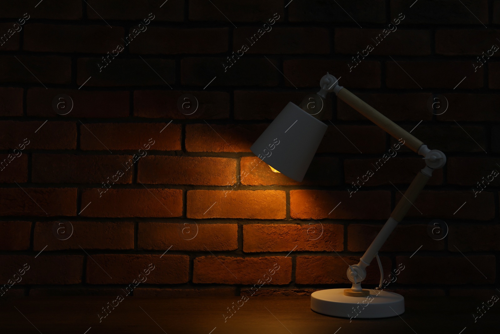 Photo of Stylish modern desk lamp on wooden table near brick wall at night, space for text