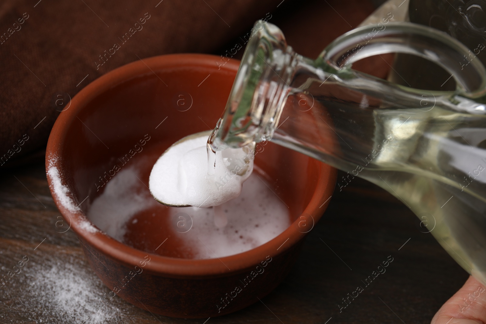 Photo of Pouring vinegar into spoon with baking soda over bowl at wooden table, closeup