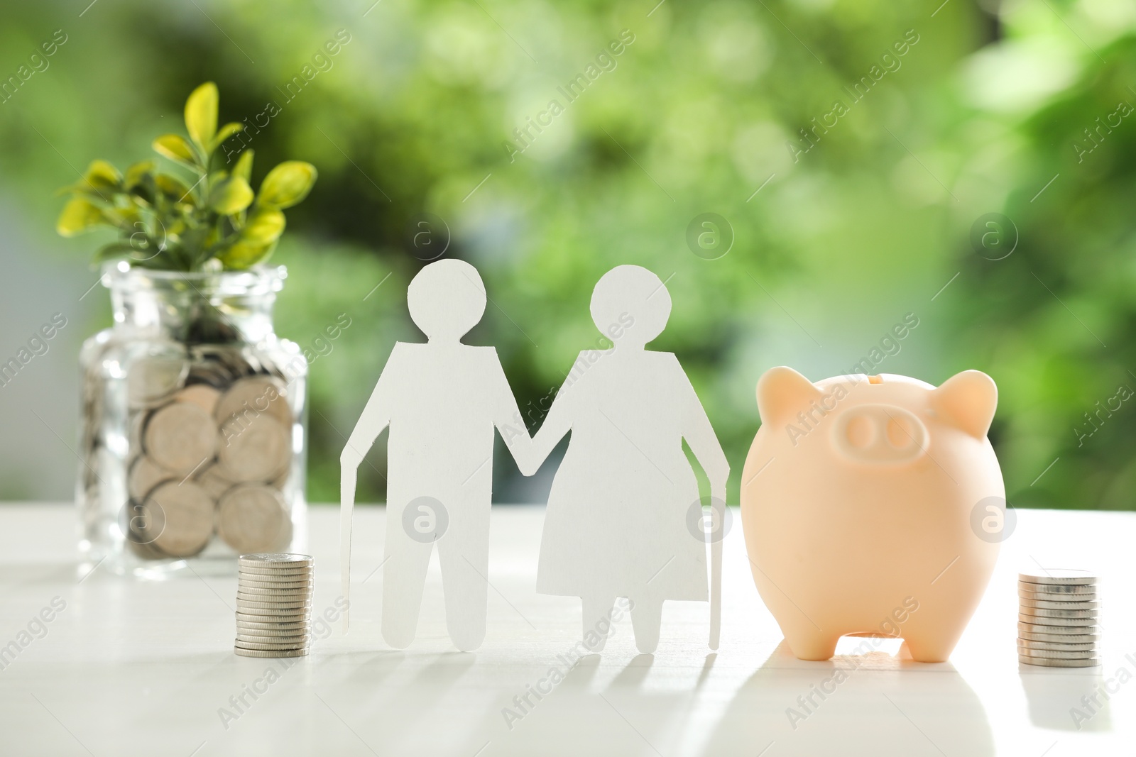 Photo of Pension savings. Figure of senior couple, piggy bank, jar with coins and twigs on white table against blurred green background