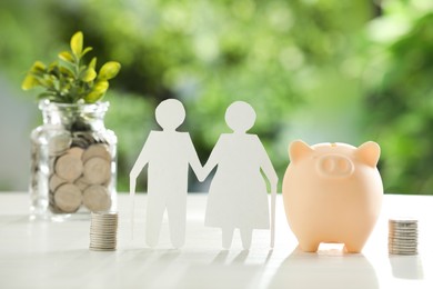 Pension savings. Figure of senior couple, piggy bank, jar with coins and twigs on white table against blurred green background