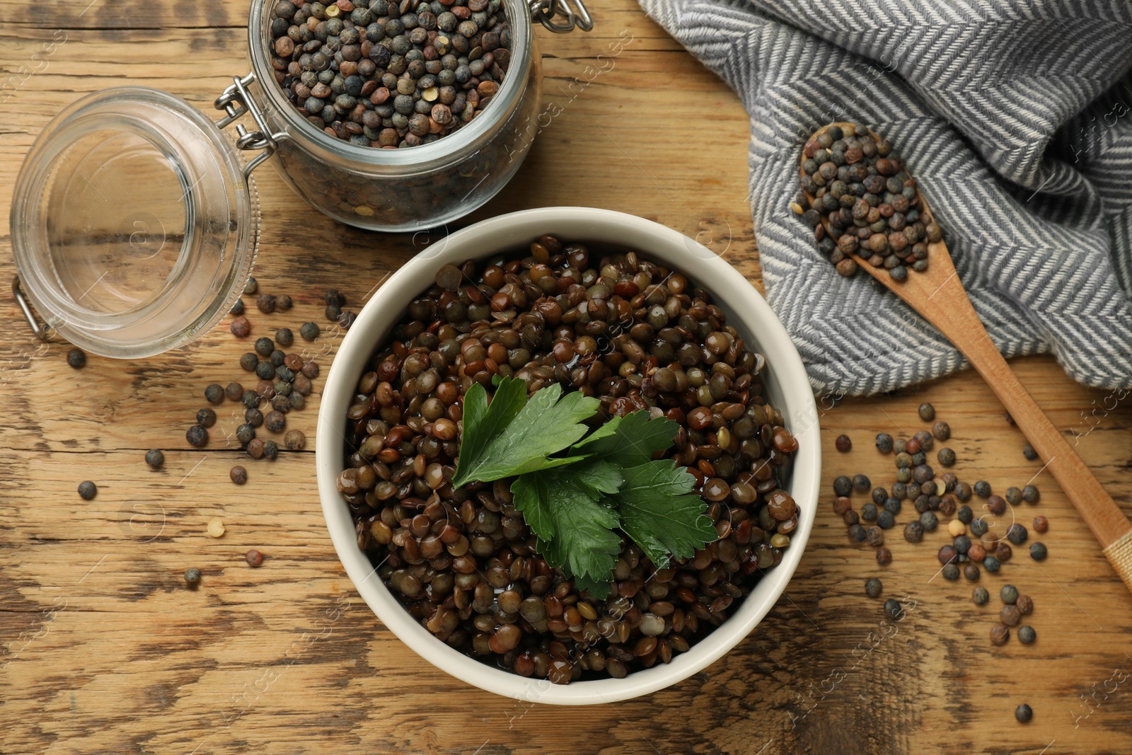 Photo of Bowl of delicious lentils, jar and spoon with raw seeds on wooden table, flat lay