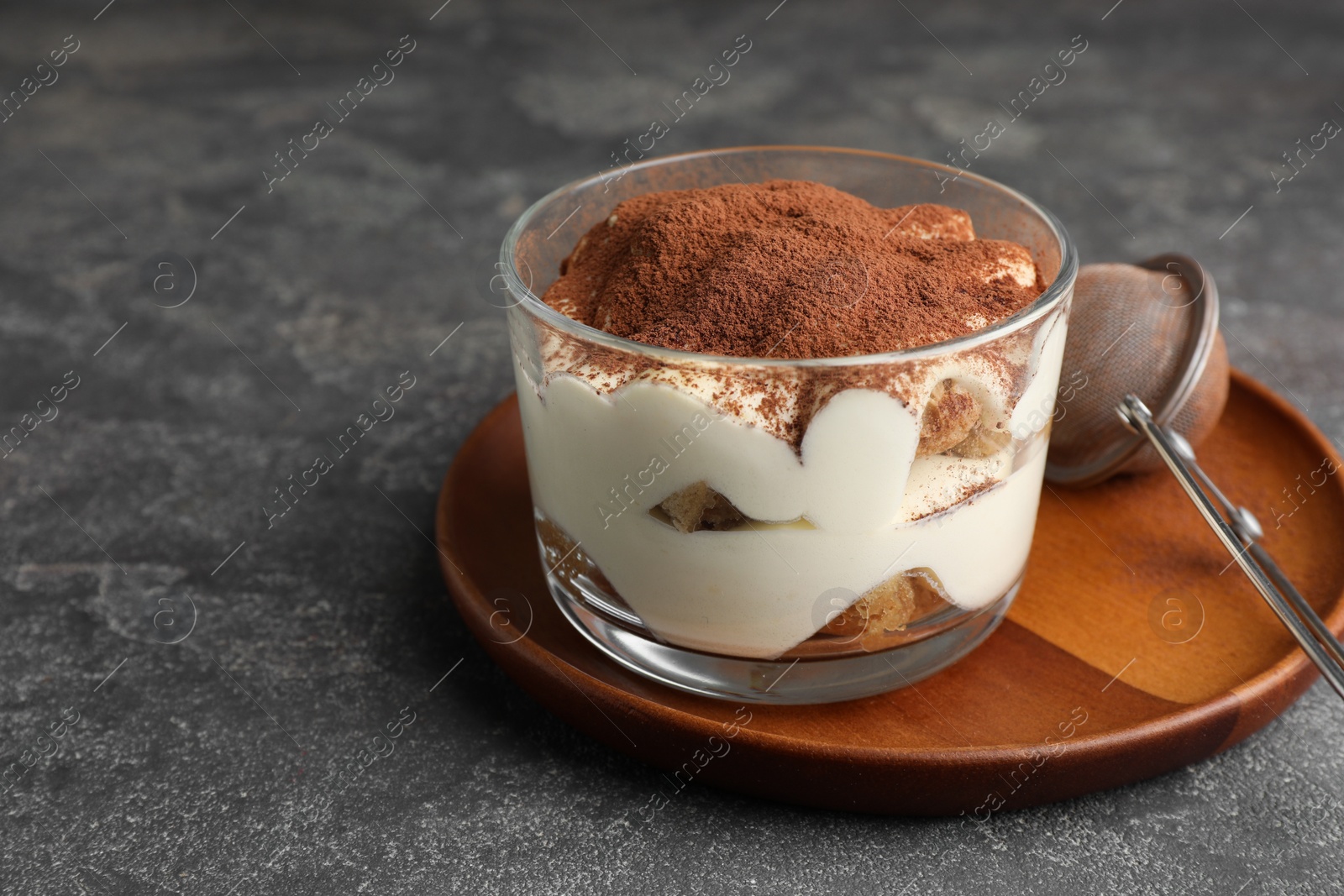 Photo of Delicious tiramisu in glass and sieve with cocoa powder on grey table. Space for text