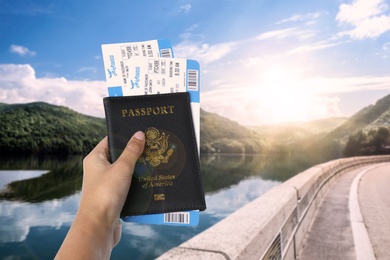 Woman holding passport with tickets near lake and mountains, closeup. Travel agency service