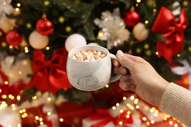 Photo of Woman holding cup of delicious cocoa with marshmallows near Christmas tree and gifts, closeup