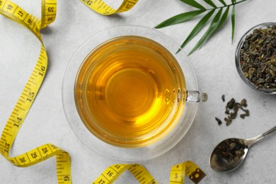 Cup of herbal diet tea and measuring tape on white table, flat lay. Weight loss concept