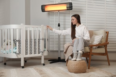 Photo of Young woman near crib and modern electric infrared heater indoors