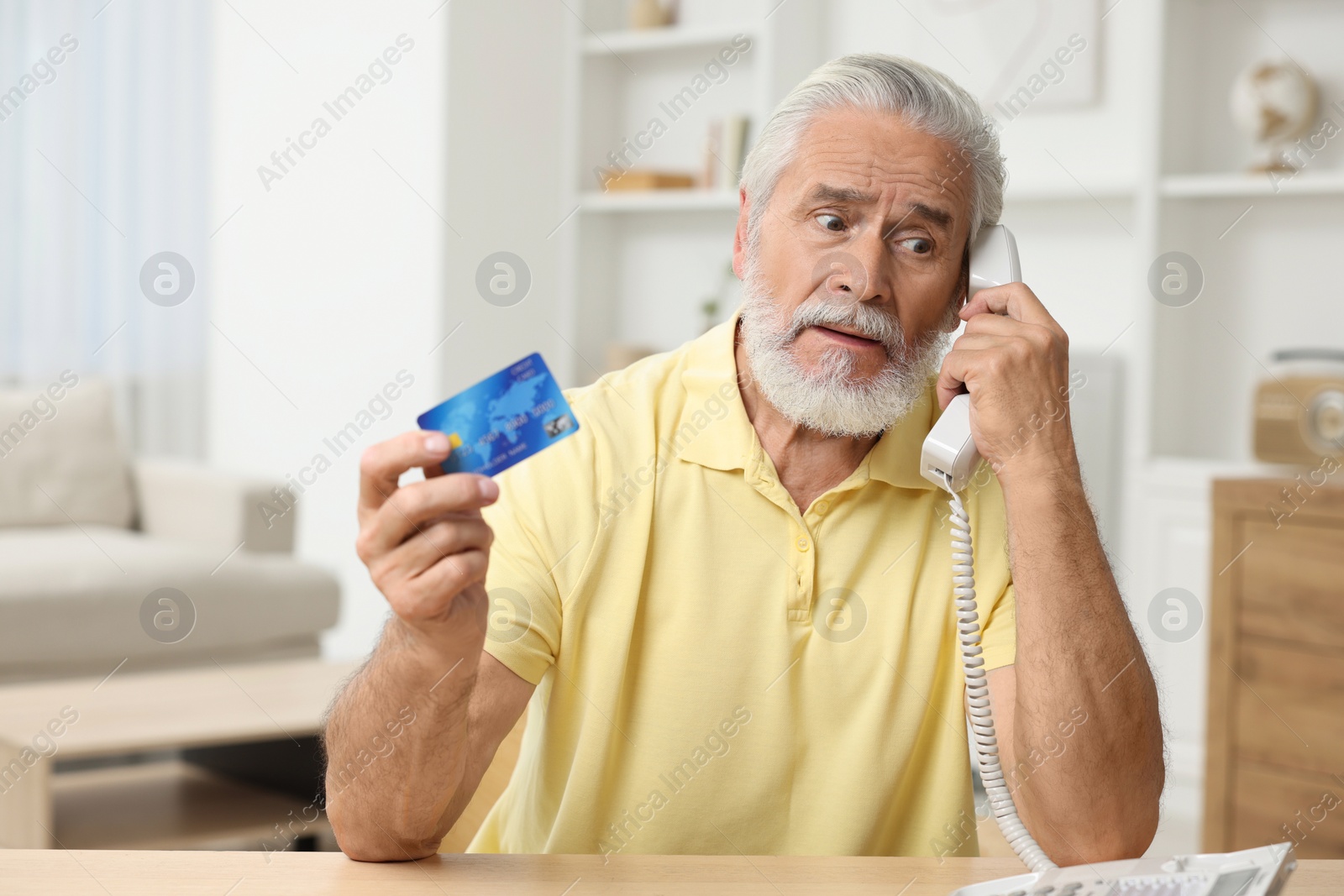 Photo of Confused senior man with credit card talking on phone at home. Be careful - fraud