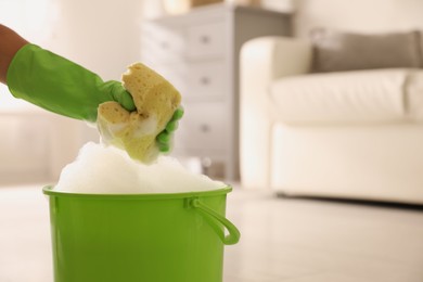 Photo of Woman holding sponge with foam over bucket indoors, closeup. Cleaning supplies