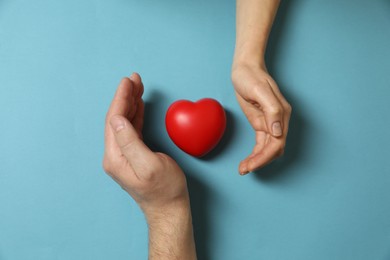 Couple protecting red decorative heart on light blue background, top view