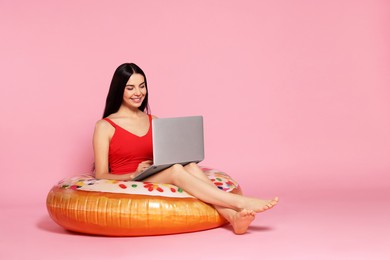 Young woman using laptop while sitting on inflatable ring against pink background