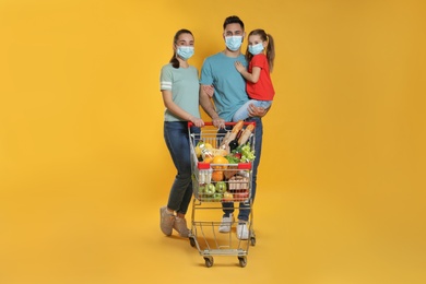 Family with protective masks and shopping cart full of groceries on yellow background