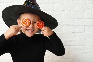 Photo of Cute little girl with candies wearing Halloween costume near white brick wall. Space for text