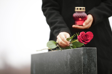 Woman with candle near black granite tombstone outdoors, focus on red rose. Funeral ceremony