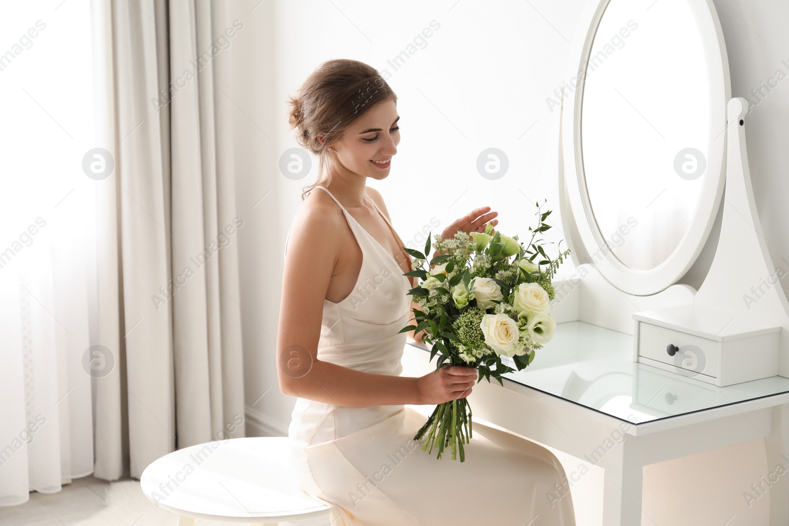 Photo of Young bride in wedding dress with beautiful bouquet near mirror indoors