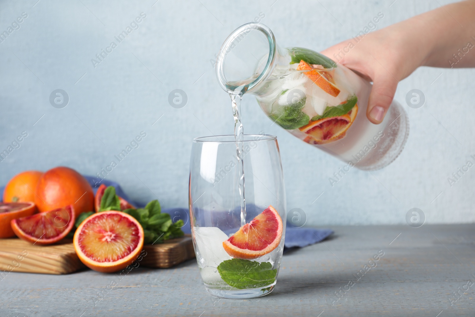 Photo of Woman pouring refreshing drink with sicilian orange from bottle into glass at grey wooden table, closeup