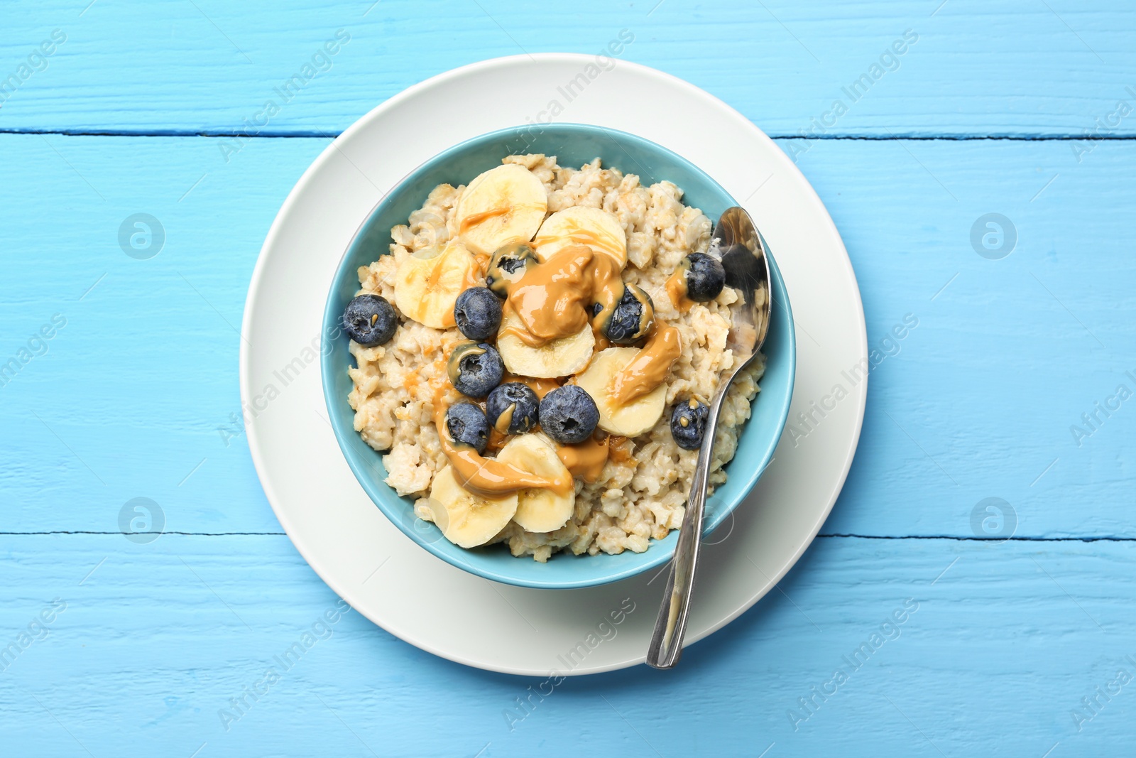 Photo of Tasty oatmeal with banana, blueberries and peanut butter served in bowl on light blue wooden table, top view