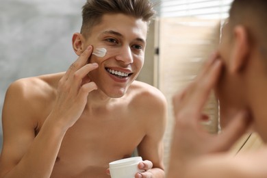 Photo of Handsome man applying moisturizing cream onto his face in bathroom