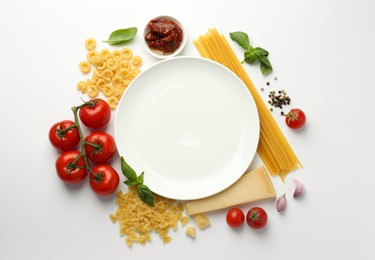 Plate surrounded by different types of pasta, products and peppercorns on white background, flat lay. Space for text