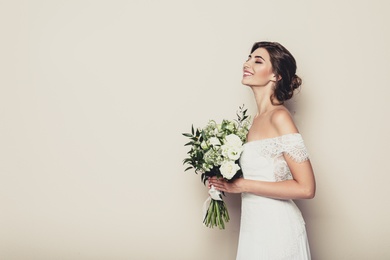 Young bride wearing wedding dress with beautiful bouquet on beige background. Space for text