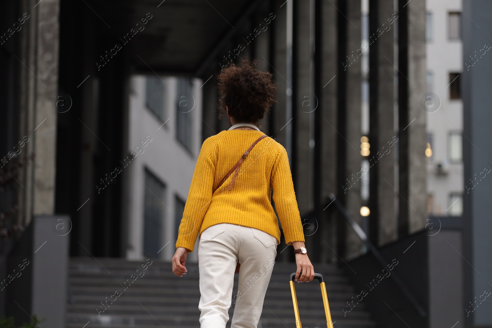 Photo of Being late. Woman with suitcase running towards building outdoors, back view