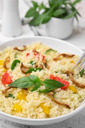 Photo of Bowl of delicious couscous with vegetables and basil served on white wooden table, closeup