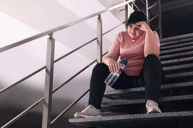 Photo of Tired overweight mature woman with bottle of water sitting on stairs indoors. Space for text