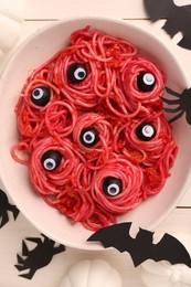 Photo of Red pasta with decorative eyes and olives in bowl on white wooden table, flat lay. Halloween food