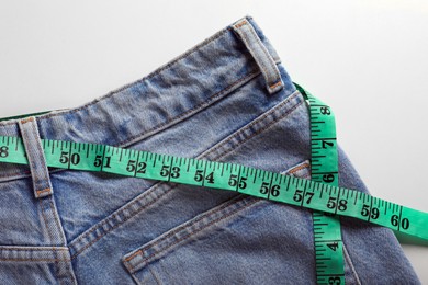 Jeans with measuring tape on light grey background, top view. Weight loss concept