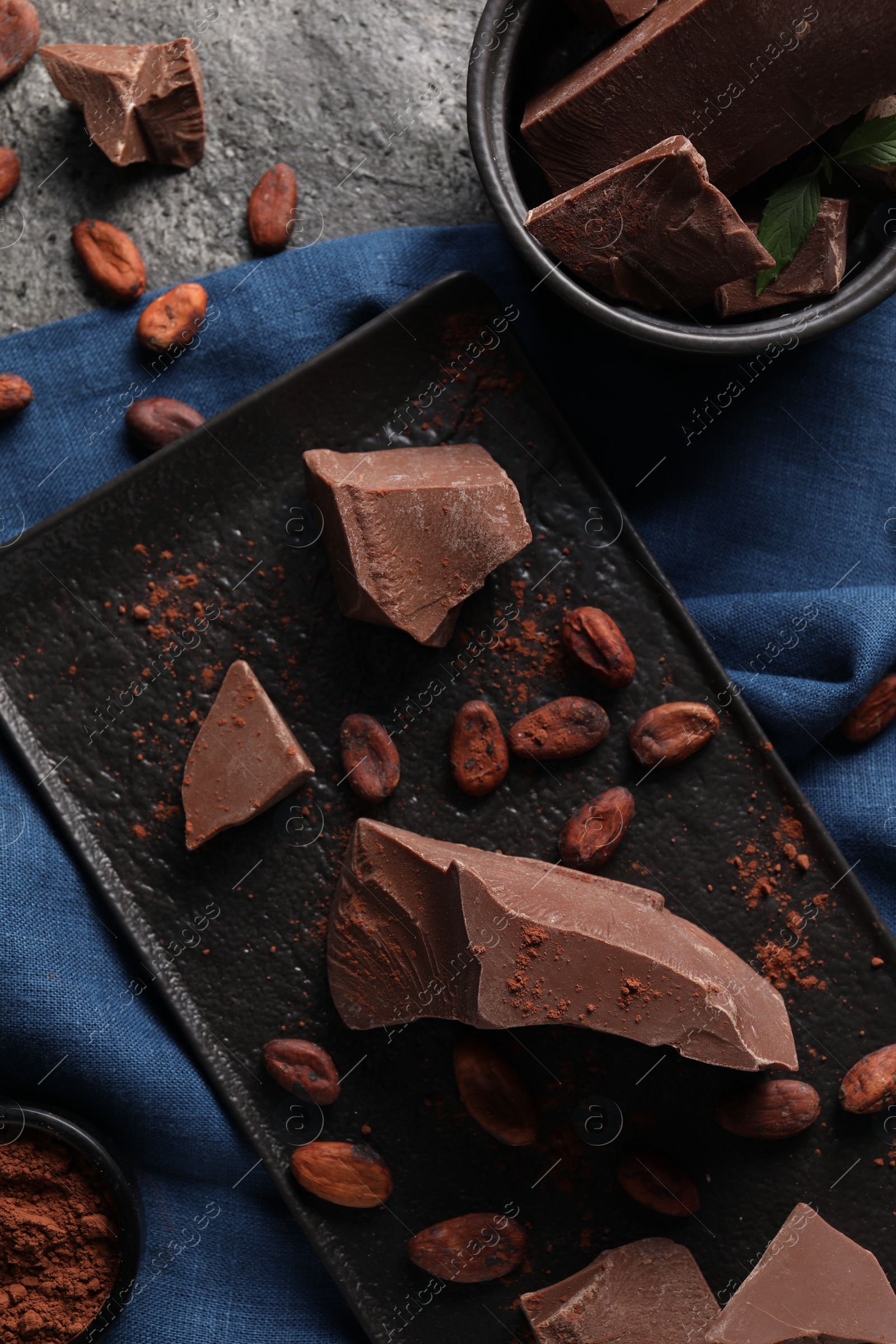 Photo of Pieces of tasty milk chocolate, cocoa beans and powder on grey table, top view