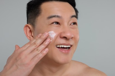 Handsome man applying cream onto his face on light grey background, closeup