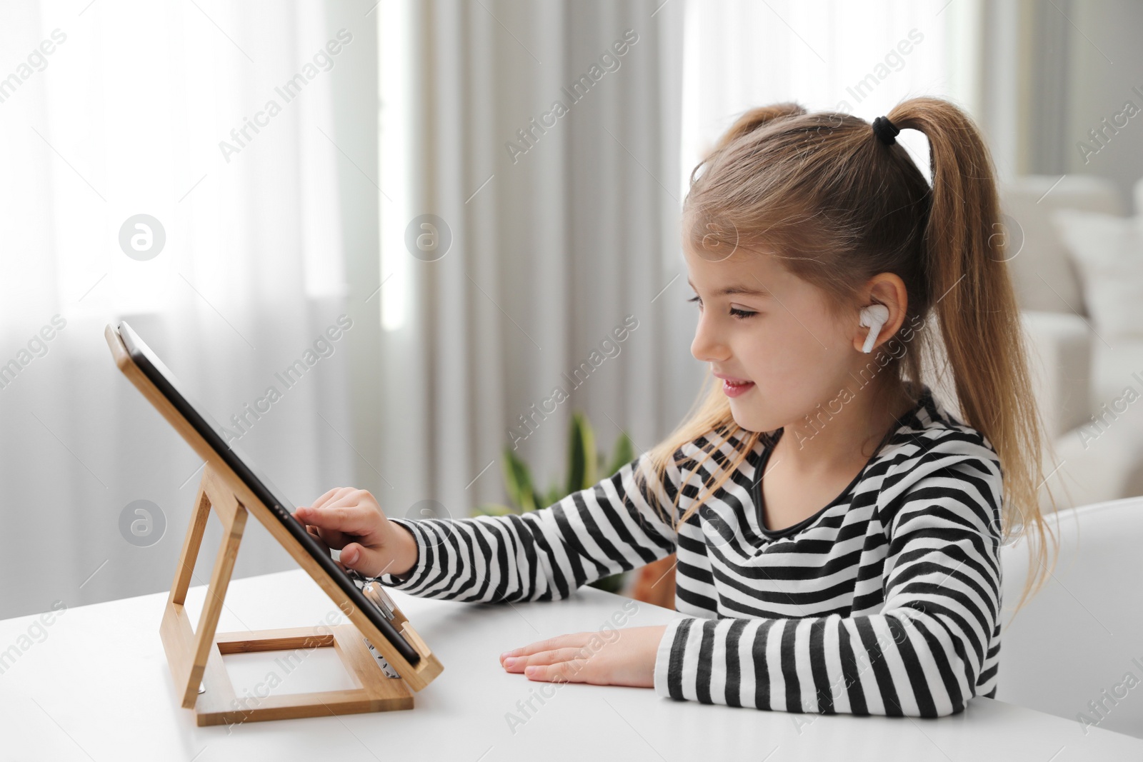 Photo of Adorable little girl doing homework with tablet and earphones at table indoors