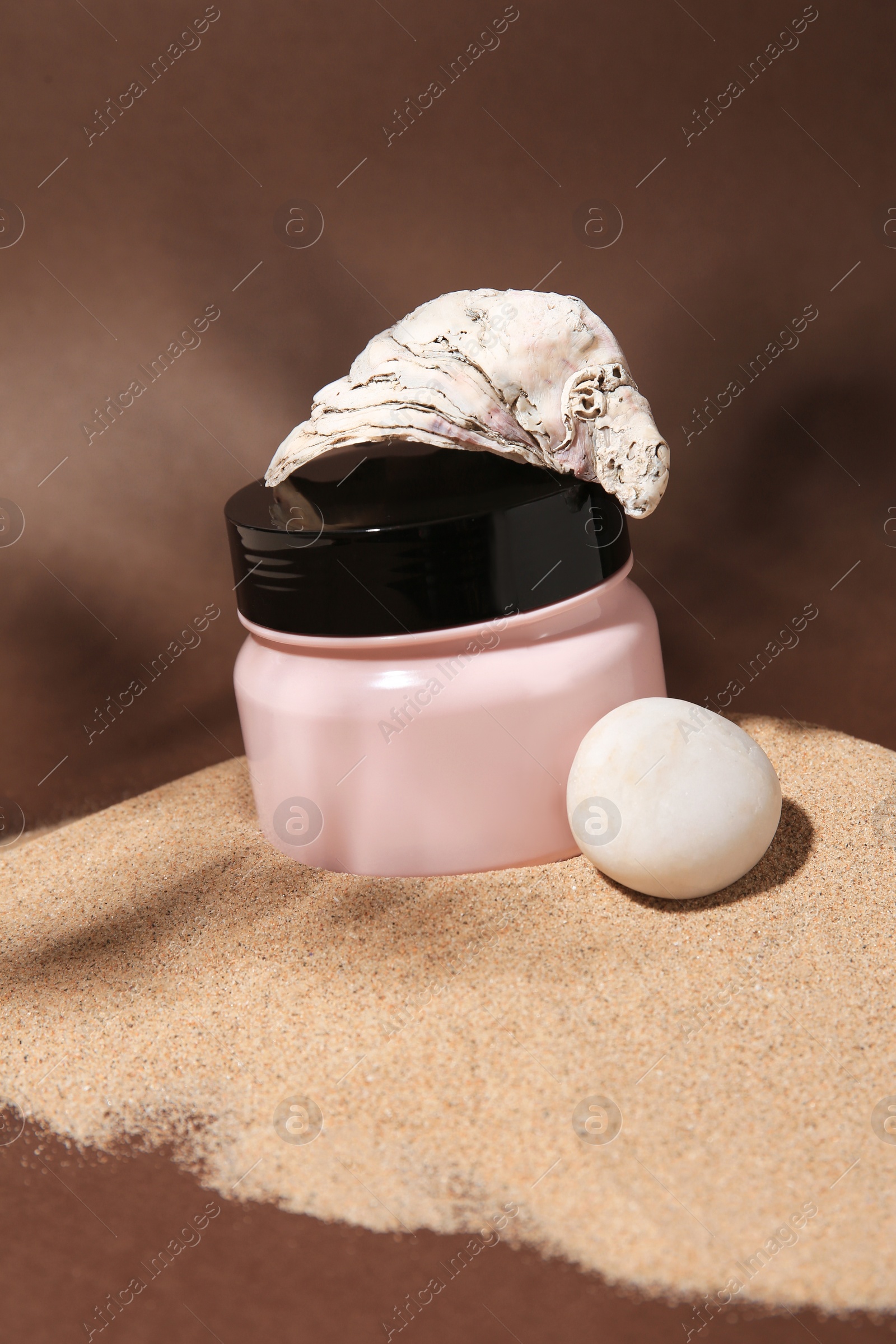 Photo of Jar of body cream, seashell and stone on sand against brown background