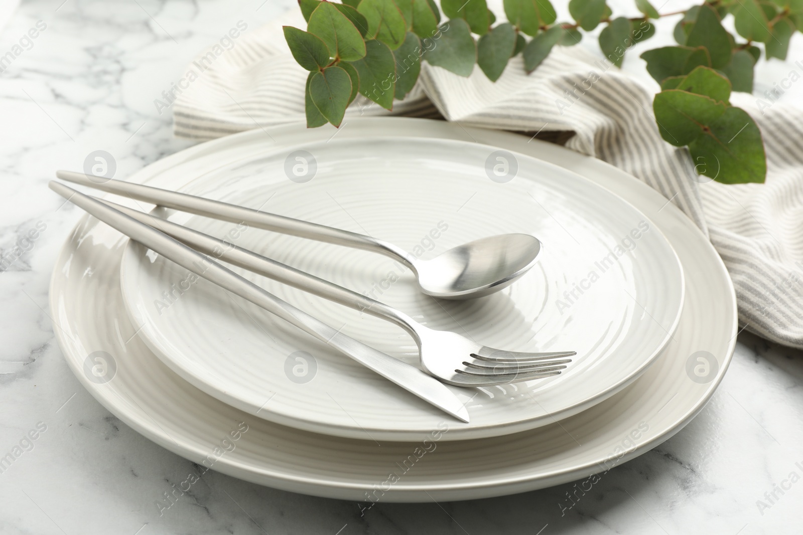 Photo of Stylish setting with cutlery, napkin, eucalyptus branches and plates on white marble table, closeup