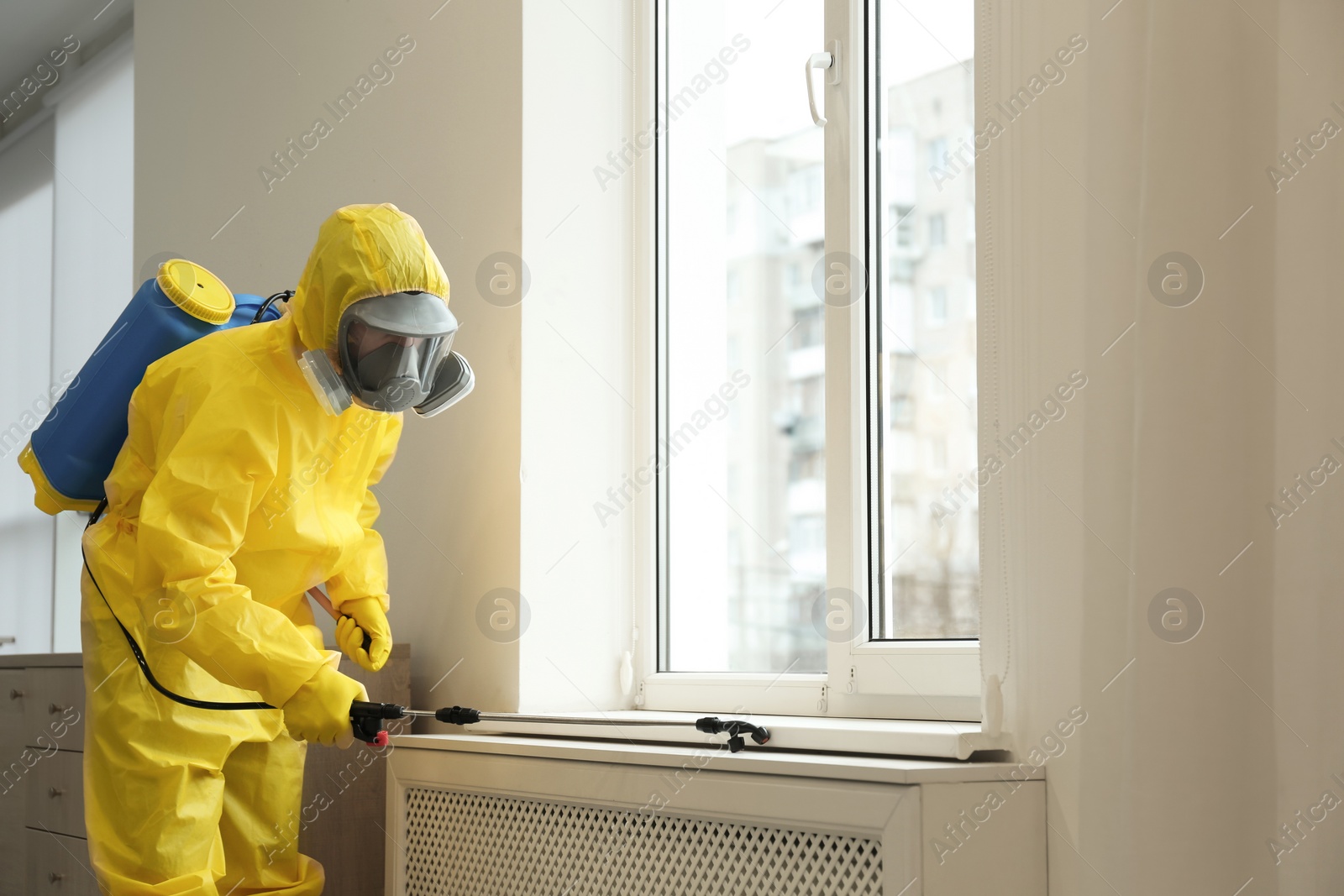 Photo of Pest control worker in protective suit spraying pesticide near window indoors