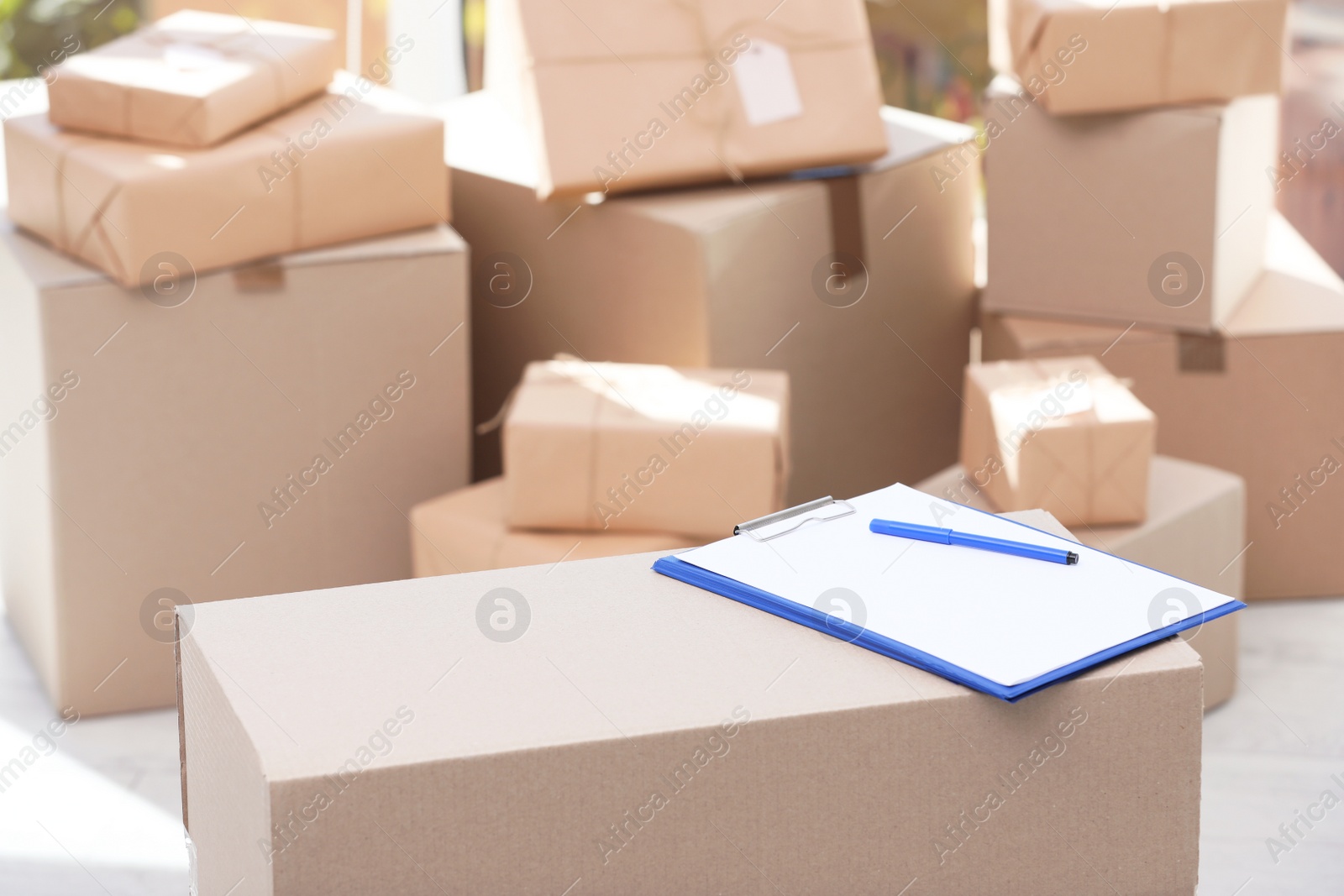 Photo of Clipboard, cardboard box and blurred stacked parcels on background, indoors