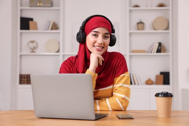 Photo of Muslim woman in hijab with headphones using laptop at wooden table in room