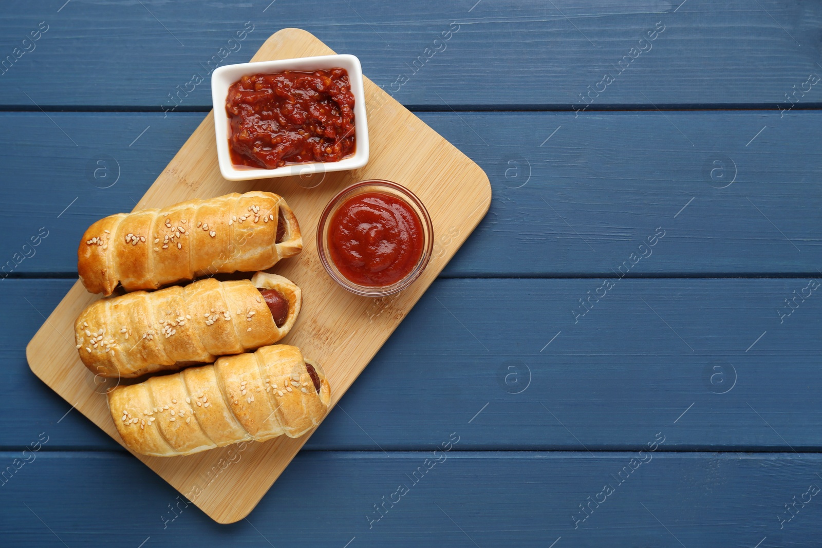 Photo of Delicious sausage rolls and ketchup on blue wooden table, top view. Space for text