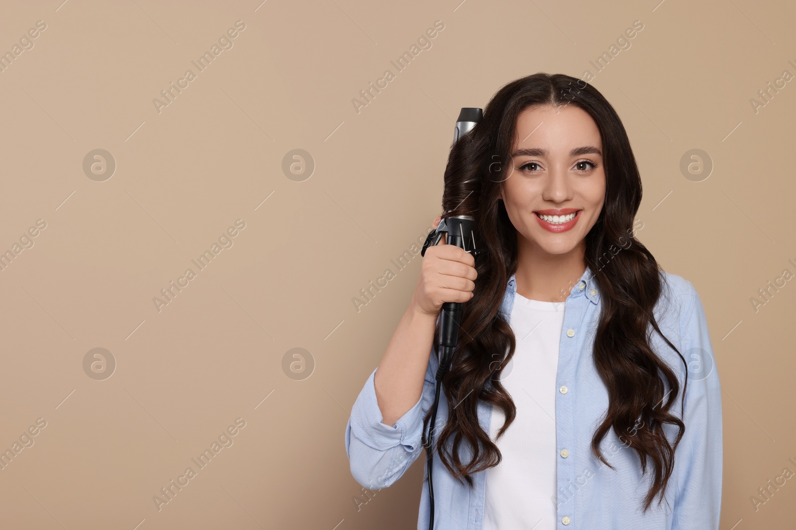 Photo of Happy woman using curling hair iron on beige background. Space for text