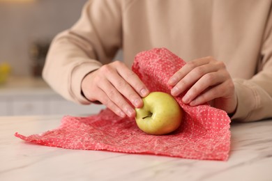 Man packing fresh apple into beeswax food wrap at light marble table indoors, closeup