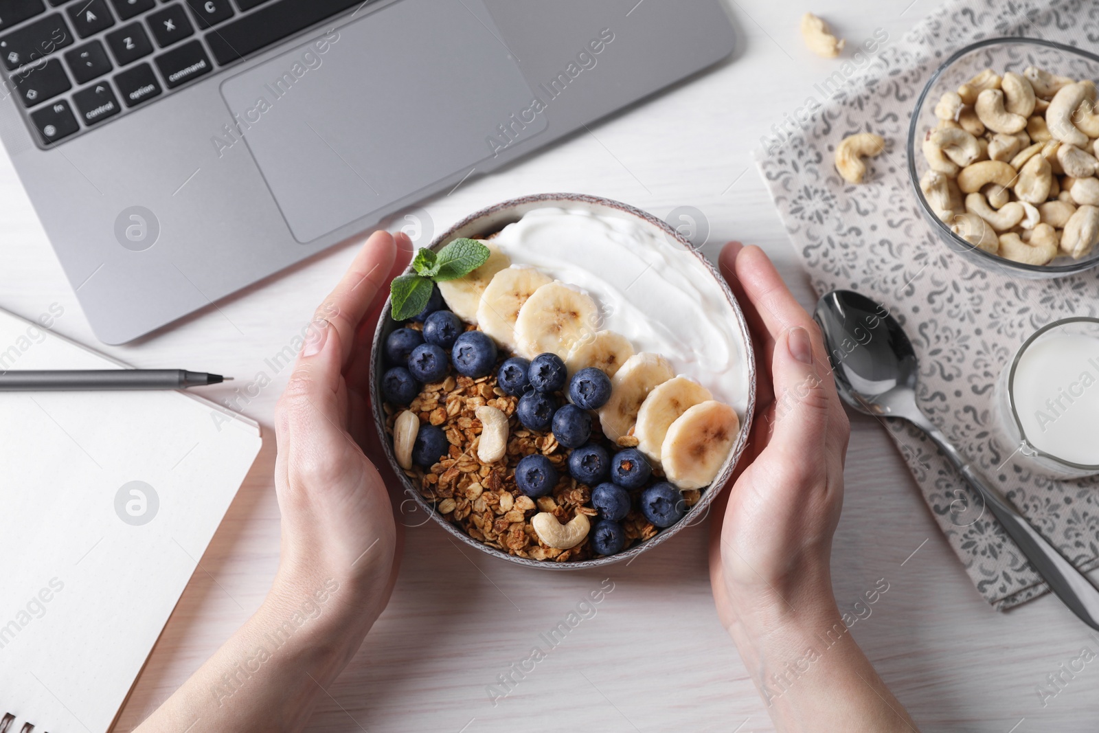 Photo of Woman holding bowl of tasty granola at white wooden table with laptop, top view