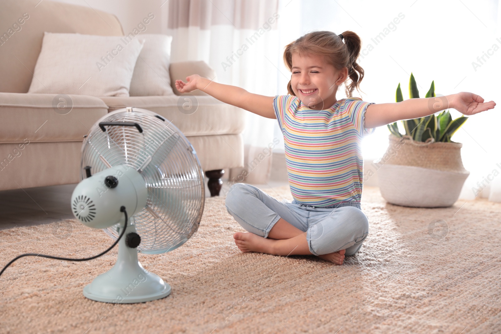 Photo of Little girl enjoying air flow from fan on floor in living room. Summer heat