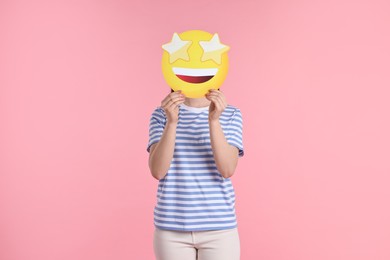 Woman holding emoticon with stars instead of eyes on pink background