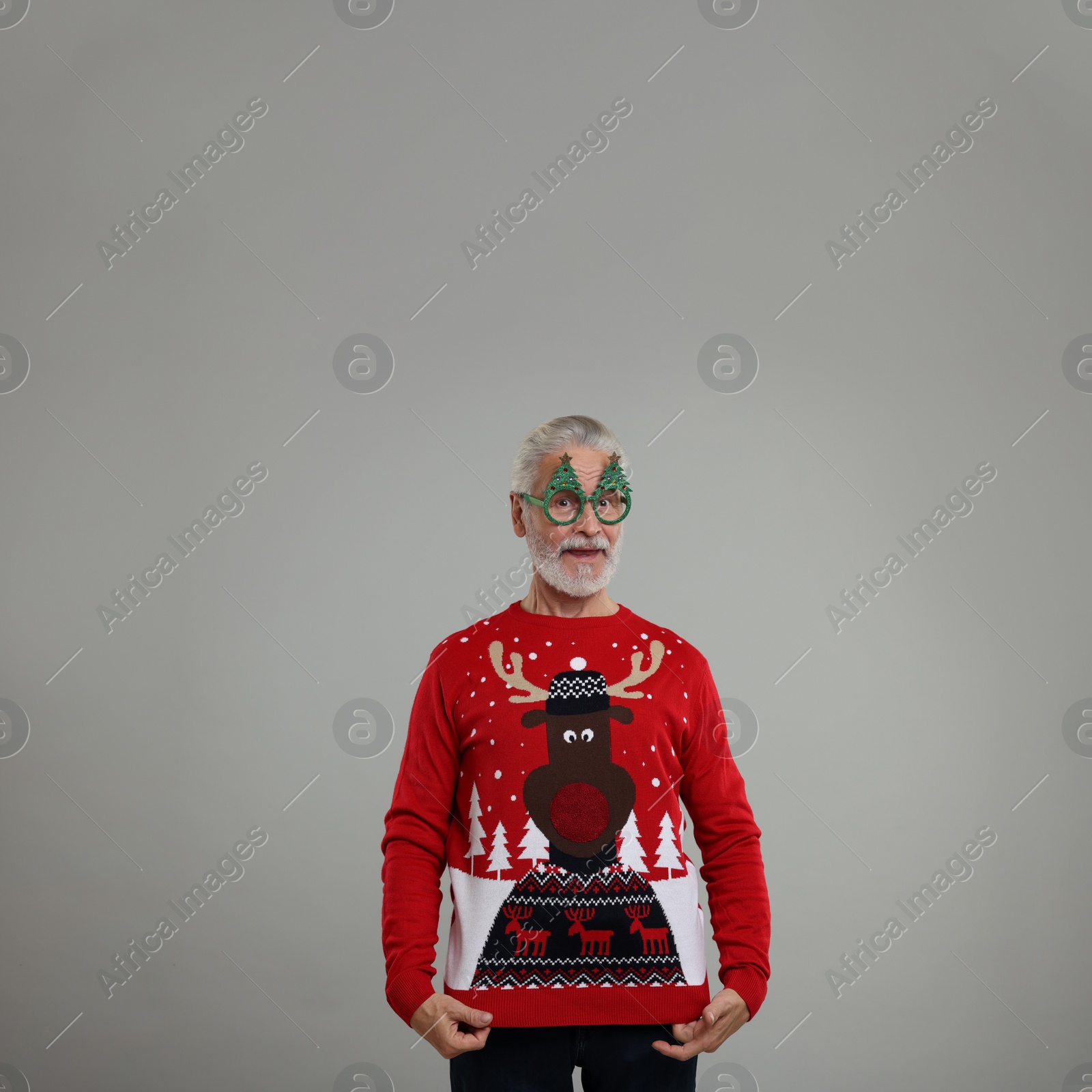 Photo of Senior man in funny glasses showing his Christmas sweater on grey background