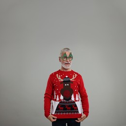 Photo of Senior man in funny glasses showing his Christmas sweater on grey background